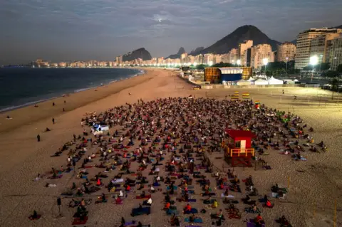 MAURO PIMENTEL/AFP An aerial view shows people taking part in a yoga event at Copacabana beach in Rio de Janeiro, Brazil, on June 22 2024. People sit on the sand of a beach with buildings and mountains in the background