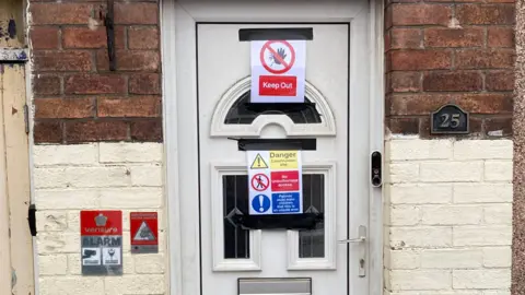 James Mountford/BBC A white front door on a terraced property which has been affixed with warning signs such as 'Keep Out' and 'Danger, Construction Site'