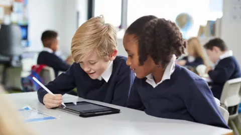 A blonde boy and a girl with black hair sit at a desk with a tablet, the boy is using a white pen to operate the tablet. They both look at the screen with interest. They are both wearing navy school uniform