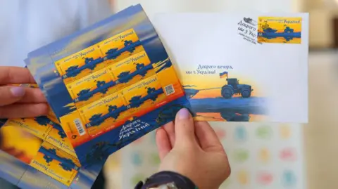 Getty Images Hand holds stamps that read "Good evening, we're from Ukraine", showing a Ukrainian tractor pulling along a Russian tank