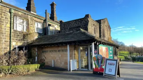 BBC A brick building with a Historic England sign and ice cream sign outside. It has glass double doors and on the side reads Abbey House Museum in green lettering. 