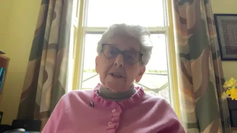 Lady Milena in pink cardigan speaking from her home