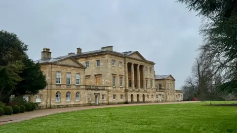 Bumble bee A large country house sits in the centre of the picture. The house has at least three floors with several pillars forming part of its front. It sits in parkland with several trees. The sky is overcast.