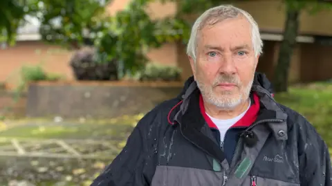 Hugh Janes in front of the registry office building. He has short grey hair and a short grey beard. He is wearing a grey raincoat and a blue and white fleece. 