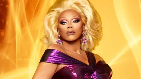 Drag royalty RuPaul is looking directly down the camera lens. She's wearing a dark purple, glittery gown with straps over her shoulders and star-shaped gold and purple earrings. Her eye make-up is dark and dramatic and she has glamorous blonde hair. She's standing in front of a background of billowing orange flames.