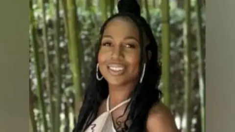 Michelle Sadio smiles at camera wearing large white hooped earrings and a pale coloured dress, in front of a green background.