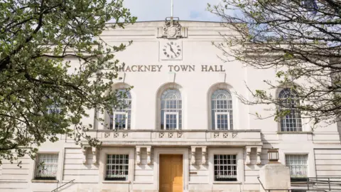 The facade of Hackney Town Hall, a cream-colored stone building with large arched windows and a clock above the entrance. Steps lead up to the main door, and trees frame the image, partially covering the building.