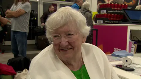 Woman with white hair wearing gold rimmed glasses, a white cardigan and a green top 