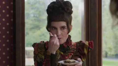 Mark Mainz Actress Shirley Henderson is dressed in a period costume. She has dark hair which is styled on top of her head. She is holding a slice of cake on a saucer and taking a bite of it from a fork. She is sitting in front of two windows and greenery from a garden can be seen in the background. 