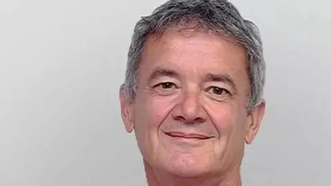 BBC Tony Dixon smiling  with short grey hair and brown eyes with grey background.