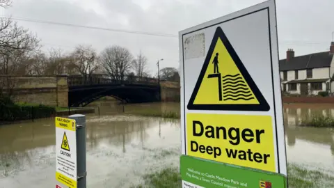 Ben Schofield/BBC A sign saying 'Danger deep water' with a river and bridge in the background. A smaller sign on the left of the image explains the site is a water meadow that is designed to flood each year. The ground behind the signs is clearly flooded and is almost indistinguishable from the channel of the river.