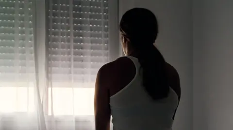 A woman with her back to the camera sits on a bed looking towards a window