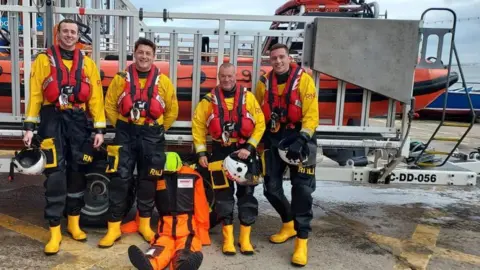 Filey Lifeboat crew members standing with the dummy in front of an orange-coloured boat wearing yellow and black clothing, with red life jackets 