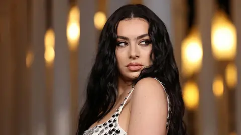 Charli XCX wearing a white dress with shiny tear drop sequins on. She is facing the camera but looking to her left away from the lens. She has long black hair and make up on