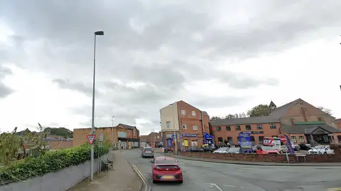 Google A road junction that bends to the right. Red brick buildings in the background, with a low wall in front of a car park. There is a lamp-post to the left and a sign that reads "give way". 