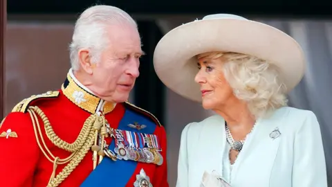 King Charles, on the left wearing a red uniform with medals attached, faces Queen Camilla, wearing a light blue jacket and a beige hat. She is also looking at him