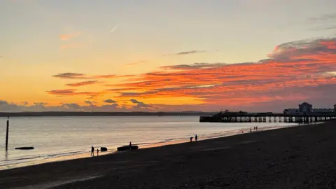 Lynn A golden sunset casts an orange and yellow glow across the sky over a beach with a pier sticking out into the water. Several people are walking along the beach.