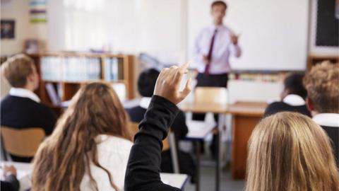 A teacher teaching pupils in a classroom in front of bookshelves and a whiteboard. In the foreground, a girl has her hand up 