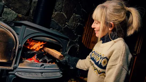 A woman putting a log into a glowing wood burning stove. She has blonde hair pulled back in a ponytail and wears a light brown jumper with a wavy pattern of blue across the front.