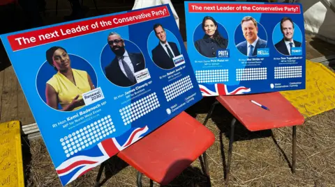 Two blue boards resting on chairs with names and pictures of the six Conservative leadership candidates