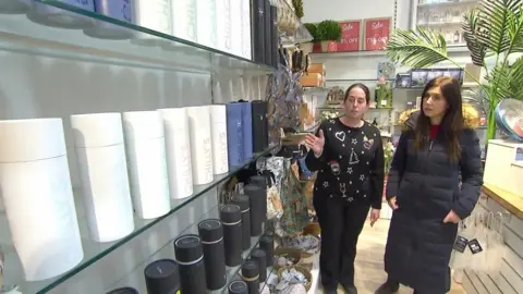 A member of staff points to a display of reusable water bottles mounted on the wall of a store.