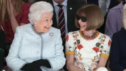 Getty Images Queen Elizabeth II and Anna Wintour sitting next to each other and smiling at a fashion show in london in 2018