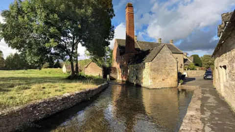 Google The Old Mill, a quaint brick building with a tall chimney and wooden paddle mill. It is positioned right next to the water on a calm sunny day