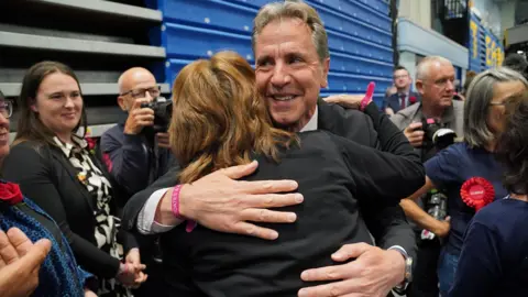 PA Media Dan Norris hugging a woman, surrounded by journalists and Labour supporters