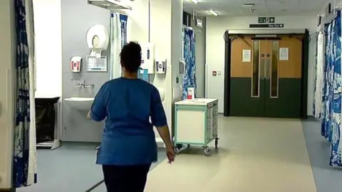 Female nurse with back to camera, in a blue uniform top and dark trousers, in an Aberdeen hospital ward, with swing doors at the end of the corridor, lined with equipment.