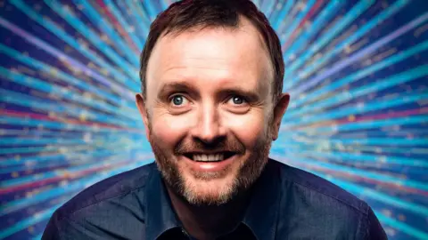 Close-up of Chris McCausland looking into the camera with multi-coloured Strictly branding in the background