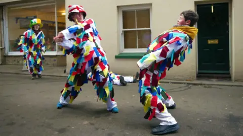 A young girl and a boy wearing white outfits covered in multi-coloured ribbons having a mock sword fight in a village street. The girl, who is wearing a hat with the English flag on it, is swinging the sword and the boy is ducking backwards. A third person in similar dress, with a hat with the Irish flag on it, cheers them on in the background.