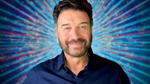 Nick Knowles, a man with dark hair and stubble, wears a dark blue shirt and stands against a blue starburst background, smiling 