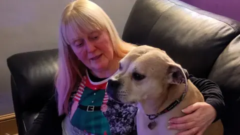 RSPCA Sue Fenn, with long blonde-white hair and wearing a Christmas elf jumper with green checks and black belt printed on it, cuddles her brown and white-haired dog Skylar on a sofa