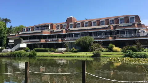 A view of the hotel from its garden. There is a lake in the foreground with the three-storey building behind it. 