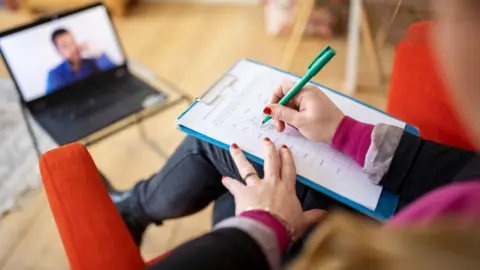 Stock photo close-up of a female mental health professional taking notes during an online cognitive behavioral therapy session with a male patient seen on a laptop.