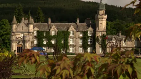 Balmoral Castle on an overcast day. It is covered in ivy and sits against a backdrop of dense forest. Green grass surounds it.