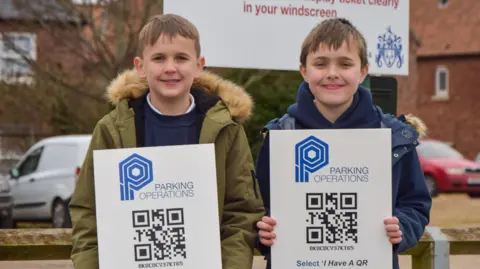 Harvey, 7,  and Arthur, 8, stand at Boal Quay car park holding signs showing how parents can use the car parking machines to get 30 free minutes parking at pick up and drop of times. 