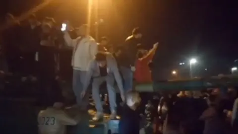 People on top of a tank, some with their arms in the air and some with their phones to hand