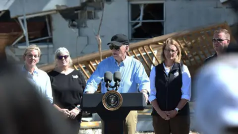 President Joe Biden speaks in Tampa, Florida after an aerial assessment of Hurricane Milton's damage - he is standing at a lectern with the US presidential stamp on it. He is wearing a black baseball cap and glasses, with a destroyed building visible behind him. 