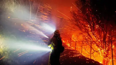 A firefighter seen at night with sparks flying off burning trees on Monday