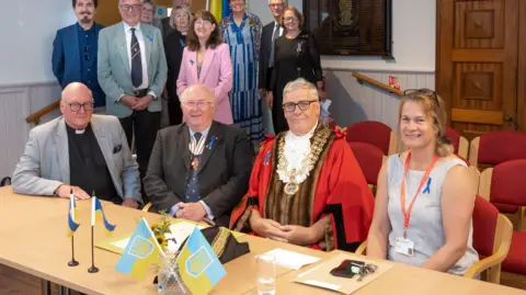 Four people sitting at a table in formal dress with small Ukrainian flags out with a small group behind them, all posing for the camera