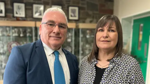 Simon and Barbara pictured in front of school awards cabinet. Simon has short white hair and is wearing thin framed dark glasses. He is wearing a dark blue suit with a white shirt and light blue tie. Barbara has short brown hair with a fringe and is wearing a patterned black and cream shirt with a black top underneath. She is also wearing a gold and silver necklace. 