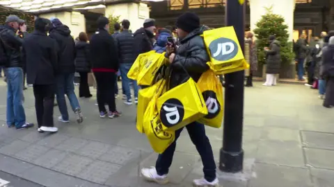 A man with several shopping bags from JD sports walking past a Boxing Day queue in central London's shopping district.