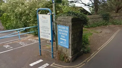 Google A blue entrance sign to the Shoalstone Seawater Pool with a white arrow pointing left towards a car park. The sign is on a concrete pillar.