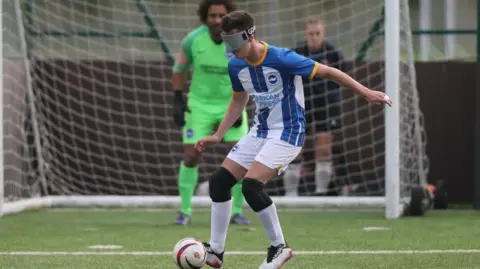 A man wearing blue and white football gear playing for Brighton in the National Blind League. He is wearing the blind football blindfold and black knee pads. He is controlling the ball, which is white with red stripes. In the background, there is a goal keeper wearing bright green gear stood in the goal post. He is also wearing black gloves. They are playing on a grass pitch, where white lines can be seen.