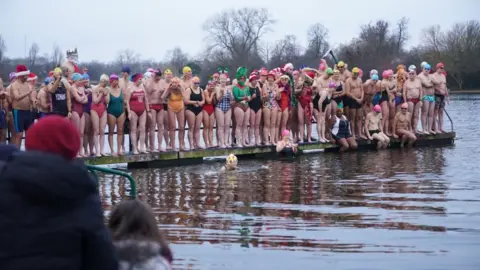 PA Media Dozens of people in swimming costumes - some in fancy dress - stand on a pontoon ready to enter a lake