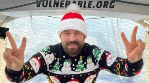 A man with a red and white hat and a Christmas sweater shows a V for victory sign with both hands.