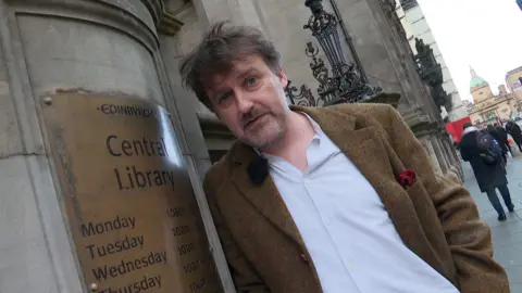 Dr Peter Mackay in a light shirt and tweed jacket, leaning on a pillar of the Central Library in Edinburgh. He is looking at the camera