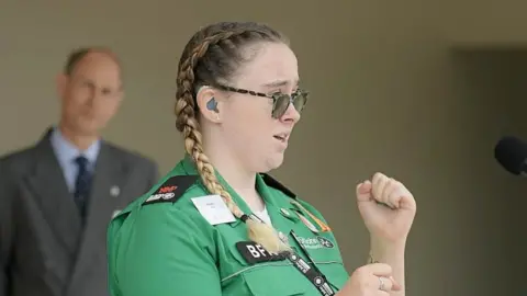 DoE/PA Media Hafwen Clarke wearing a green St John Ambulance shirt and her blonde hair is in pigtails, her hands are up as she is signing