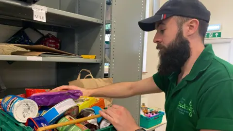 A man is to the right of the image, he is wearing a green t-shirt which says "foodbank" on it. He has a blue cap on, and also has a dark brown beard. He is picking up a crate of tinned food and cans in a food bank.